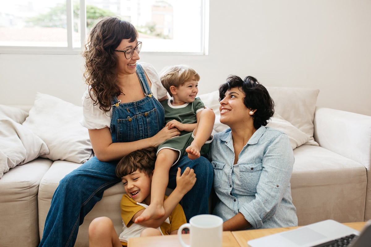 Two women and two young children laughing together on a couch in a bright living room.