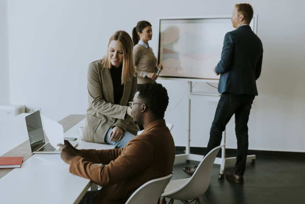 Four colleagues collaborate in an office, two at a desk with a laptop and two near a presentation board.
