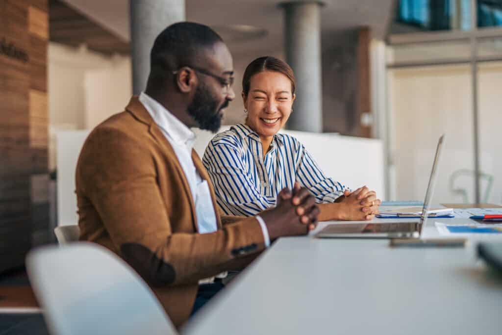 Two colleagues smiling and sitting at a desk with a laptop in a modern office setting.