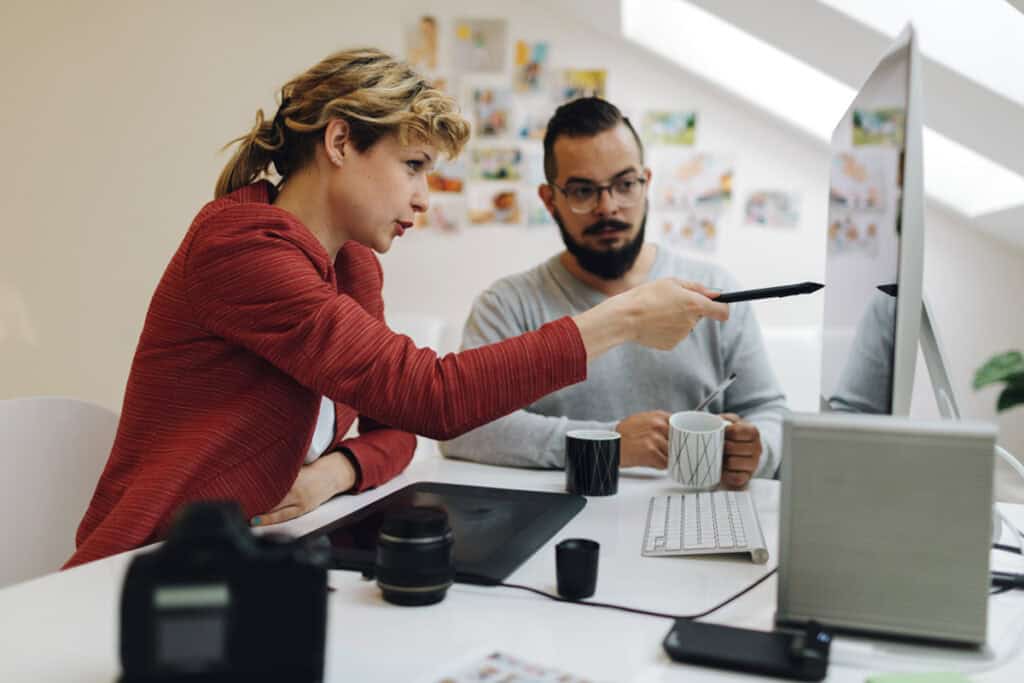 Woman pointing at computer screen while man watches; both seated at desk with coffee cups and camera equipment.