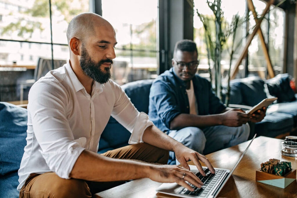 Two men sitting indoors; one types on a laptop while the other reads a book on a couch in the background.